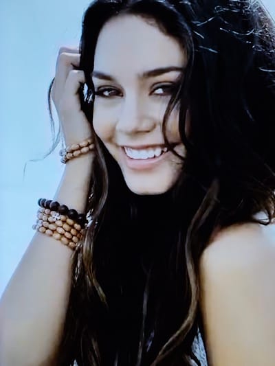 The media file is a close-up, rotated photograph of a young woman with long, dark hair. She is lying down, with her head tilted and her face partially obscured by her hair and arm. Her eyes are open and looking towards the camera, and she has a slight smile on her lips. She is wearing two bracelets on her left wrist, one made of light-colored wooden beads and the other of darker beads, both with small metallic charms. The background appears to be a light-colored, possibly textured surface, and a bright, light blue area which might represent a wall or fabric. The lighting suggests an indoor setting, possibly a studio or a bedroom, with soft illumination. The overall mood of the image is relaxed and intimate. There is no visible text. The image is taken in Švenčionys, Lithuania.
