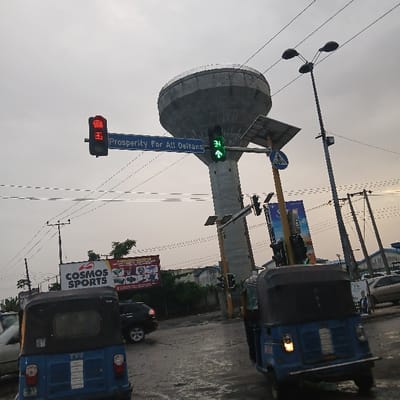 The image displays an urban street intersection in Warri South, Delta State, Nigeria, under an overcast sky. The road surface is wet. A large, grey, concrete water tower with a circular upper tank structure is prominent in the upper center. To the left, a red traffic light with a digital countdown showing '3' is mounted above a blue sign that reads "Prosperity For All Deltans." To the right, another traffic light displays a green upward arrow with a digital countdown showing '34'.

In the foreground, two blue auto-rickshaws (Keke Napep) are visible; the one on the left has "TVS" on its rear, and the one on the right has its headlights illuminated. A dark-colored SUV is partially visible in the mid-ground behind the left auto-rickshaw. Further in the background, a light-colored sedan and other vehicles are present. A large billboard on the left displays "COSMOS SPORTS," and another billboard is partially visible on the right. A blue pedestrian crossing sign is visible on a pole to the right of the water tower. Numerous utility poles and overhead wires are present throughout the scene. One individual in a white shirt is partially visible standing on the far right side of the image.
