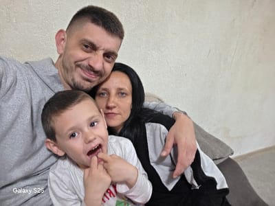 The image depicts three Caucasian individuals – an adult male, an adult female, and a young male child – positioned closely together indoors in Karnalovo, Bulgaria. The adult male, with short dark hair and a beard, is smiling broadly, wearing a grey hooded sweatshirt, and has his left arm around the adult female's shoulders. The adult female, with long dark hair, wears a grey and black garment and maintains a neutral expression, her right hand resting on the male's arm, displaying a silver bracelet on her wrist. The young male child, with short light brown hair, is positioned in the foreground, wearing a light-colored t-shirt with a partial graphic visible. He has an open-mouthed expression and is seen touching his lower teeth with his left index and middle fingers. The background features a plain, textured, light-colored wall with minor marks and a segment of a light-toned upholstered furniture piece. The bottom left corner of the image contains a "Galaxy S25" watermark.