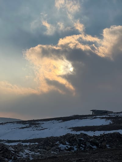 The image displays a barren, rocky landscape in Talfita, Syria, under a dramatic, partially clouded sky. The sun, a bright disc, is visible through a dense formation of yellowish-orange and grey clouds, illuminating them from behind. The upper left portion of the sky is clearer, showing a pale blue hue. The foreground and mid-ground consist of dark, rugged earth and numerous grey rock fragments, with irregular patches of white snow distributed across the undulating terrain. On a distant rise to the right, a solitary, low-profile, dark-colored structure, appearing to be constructed from robust materials, stands. It features a flat or slightly sloped roof. No individuals, animals, or discernible actions are present. The lighting conditions suggest either early morning or late afternoon.