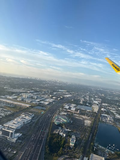 The image presents an aerial view over Dania Beach, Florida, United States of America. A portion of a yellow aircraft wing with the text "Howdy" is visible in the upper left corner. Below the wing is a clear blue sky fading to a hazy horizon. Below the horizon is a cityscape including highways, buildings, lakes, and parking areas. The scene conveys a sense of flight and perspective over a densely populated region.