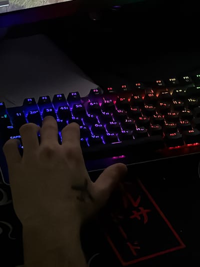 A person's hand rests on a backlit keyboard. The keyboard displays a spectrum of colors, predominantly blues and reds. The hand has a mark or drawing on it. A patterned surface lies adjacent to the keyboard, with some visible Japanese-style writing on a red-outlined section. The keyboard appears to be a gaming keyboard with function keys clearly visible.