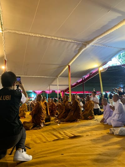 The image shows a large group of people seated on the floor under a white tent. The people are seated in a large circle, and they are all facing the center of the circle. The people in the center are dressed in saffron robes, and the people in the outer circle are dressed in a variety of clothing. The people in the center appear to be Buddhist monks. There are several people standing around the edge of the circle, including a man taking a photo with a smartphone. The floor is covered in a yellow carpet. The image was taken in the city of Mungkid, Indonesia, at the coordinates -7.605 latitude and 110.23 longitude. The tent is made of white canvas and has a white fringe running along the seams. The event is taking place at night and is lit by artificial lights. The image is lit from above by several lamps. The roof of the tent is made of white canvas, and the poles are made of metal. There are several people in the background standing around the edge of the tent. There are some trees and foliage visible in the background. The image is taken at a slightly upward angle.