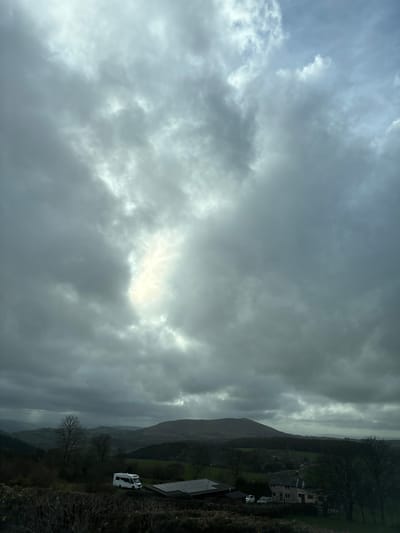 The image depicts a landscape in Shrewsbury, United Kingdom, under a cloudy sky. A prominent hill or mountain is visible in the background, partially obscured by mist or rain. Closer to the foreground, there's a building with a roof and a white van parked nearby. Some trees and foliage are present, though details are limited due to the overcast conditions. The overall scene is subdued and potentially wet due to the atmospheric conditions.