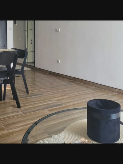 In Bielawska Wieś, Poland, an indoor scene depicts a dining area with modern furniture. A dark-colored dining table with chairs is partially visible on the left, with a glass-topped coffee table in the foreground. On the coffee table sits a dark blue fabric container and a round woven placemat. The floor is laid with wooden planks, and a light-colored rug is placed beneath the coffee table. The walls are white, with a glass-partitioned door and frame visible in the background. Light fixtures are present on the wall.