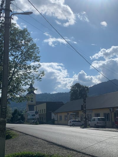 The image captures a roadside scene in Yasinia, Ukraine, under a clear blue sky with scattered cumulus clouds and direct sunlight from the upper right. In the immediate foreground on the left, a tall concrete utility pole with a streetlamp extends upwards, supporting multiple overhead power lines that traverse the frame diagonally. Behind the pole, a large deciduous tree with green foliage is partially visible.

A two-lane asphalt road with solid white lane markings occupies the foreground and midground, appearing clear of pedestrians or moving vehicles. Along the right side of the road, a row of buildings with a predominantly light yellow/cream facade and gray sloped roofs is visible. Parked curbside in front of these buildings are a light-colored station wagon and a white passenger van; a red vehicle is partially visible at the far right edge of the frame.

On the left side of the road, in the midground, stands a multi-story building featuring a prominent church-like tower. The tower is light-colored, with a dark, tiered, onion-domed spire. Adjacent to this structure is a darker building and a large billboard displaying an advertisement. The background is dominated by densely forested mountains under the bright sky. No individuals are visible in the scene.
