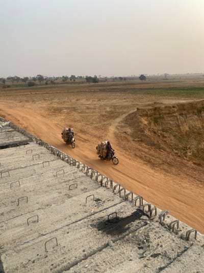 The image, when viewed in its correct upright orientation, presents a scene from a rural or undeveloped area under a hazy, pale sky.
In the foreground, running diagonally from the lower right towards the upper left, is a substantial concrete structure, possibly a wall, barrier, or part of an unfinished construction project. Its surface is rough and textured, exhibiting signs of wear or incomplete finishing. Numerous exposed rebar (reinforcing bars) protrude along its top edge and intermittently from its vertical face. These rebars form distinct U-shapes or hooks, suggesting the structure is either under construction, awaiting further reinforcement, or is a remnant of a larger edifice.
Parallel to this concrete structure and immediately adjacent to its base, is a wide dirt road or path. Two motorcycles are visible on this unpaved road, traveling from the right side of the frame towards the left. Both vehicles are heavily laden with goods. The motorcycle closer to the foreground carries large, irregularly shaped bundles, possibly wrapped in fabric, strapped securely to its rear and sides. A rider is seated atop the packed cargo. The second motorcycle, positioned slightly further back and to the right, is also heavily loaded in a similar fashion. A noticeable plume of dust trails behind this second motorcycle, indicating its movement along the dry, unpaved surface.
Beyond the dirt road, the landscape stretches far into the distance. It comprises vast, dry fields of a light brown or yellowish hue, characteristic of arid or semi-arid terrain. Sparse trees and shrubs are scattered across this expansive area, with a more concentrated line of trees visible in the mid-distance, potentially marking a boundary or a water source. The terrain appears gently undulating. A subtle line of greener vegetation is visible further back, hinting at a change in agricultural use or the presence of irrigation.
The sky occupies the upper left portion of the image and is uniformly light, almost white or pale gray, indicative of a hazy atmosphere, possibly due to dust, fog, or an overcast condition. The diffused light suggests it is daytime, likely mid-morning or afternoon, but without strong, defined shadows to pinpoint the exact time. No discernible text is visible in the image.