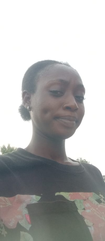 A young Black woman with dark skin, dark eyes, and dark hair pulled back into a bun is shown in a close-up, shoulder-up portrait. She is wearing a black t-shirt with a floral pattern in shades of pink, green, and white. The woman is looking towards the camera with a slight smile and dimples on her cheeks. She has a small, silver earring in her left ear. The background is a bright, overcast sky with a hint of trees visible in the lower right corner. There is no indication of the location being in Kukum, Nigeria.
