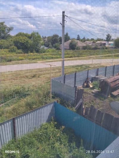 The image shows a view of a rural area with houses and vegetation. In the foreground, there is a corrugated metal fence, partly blue and partly silver. Behind it, there are some wooden planks, bricks, and a large rusty metal object resembling a tank or barrel. To the left, a road or path runs horizontally across the frame, bordered by dry grass and some green vegetation. Further back, a line of trees forms a dense green background, with a few houses visible among them. A utility pole with wires stands tall in the middle ground. The sky is partly cloudy with white clouds against a blue background. The timestamp "28.06.2026 13:47" and "REDMI 15" are visible in the bottom corners. The scene appears to be in the village of Aiyt, Kazakhstan, based on the provided context.