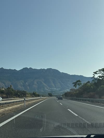 This is a landscape image captured from a vehicle driving on a highway. The highway runs parallel to a steep, rocky mountain range covered in sparse vegetation. The sky is a clear, pale blue, suggesting a sunny day. The time of day appears to be late morning or early afternoon, as indicated by the lighting.
The highway has two lanes and is marked with white lines. A guardrail separates the road from the mountainous terrain. Several vehicles are visible on the highway in the distance, including a silver car and a truck. Signs indicating speed limits or distances might be present, though they are not clearly legible. One sign with the number "130" is visible on the road surface, likely indicating a speed limit.
The mountains are rugged and show signs of erosion, with deep ravines and sharp peaks. Green foliage, likely trees and shrubs, clings to the lower slopes of the mountains and lines the side of the highway. The scene evokes a sense of travel and exploration, with the open road leading towards the majestic natural landscape. The location context of Altea, Spain, suggests a Mediterranean setting.