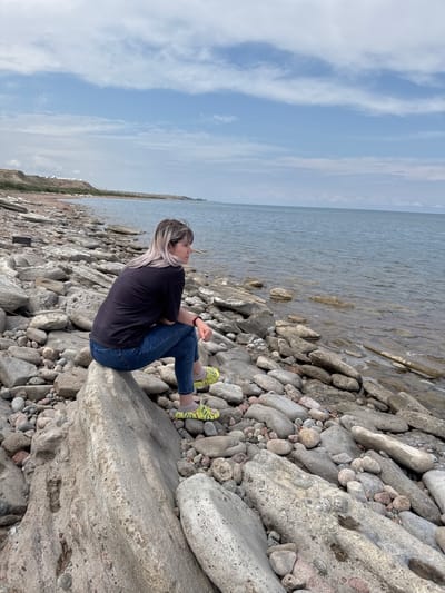 A woman with blonde hair, wearing a black t-shirt and blue jeans, sits on a large rock on a rocky shore. She is looking out at a calm body of water under a cloudy sky. The shoreline is covered in a variety of smooth, grey and pinkish rocks of different sizes. In the distance, a low, brown hill is visible along the horizon, with some white structures on its crest. The water appears clear, with small waves lapping at the shore. The overall scene is one of quiet contemplation in a natural, rugged environment. The location is the shore of Issyk-Kul Lake, near the city of Tong, Kyrgyzstan.