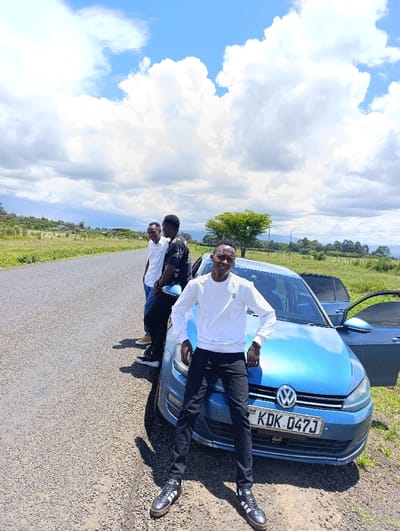 Three young men are standing next to a blue Volkswagen Golf, parked on a paved road. The man in the center is wearing a white sweatshirt, dark pants, and black sneakers. The other two men are wearing black shirts and jeans. The car has a Kenyan license plate that reads "KDK 047J". The scene is located in Njoro, Kenya at latitude -0.292 and longitude 35.9. The sky is bright blue with puffy white clouds.  There is green grass and trees in the background. The men are looking towards the camera. The car appears to be clean and in good condition. The road is empty. The environment is rural and sunny.