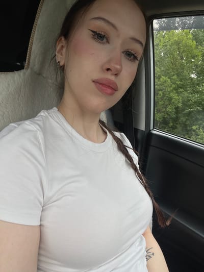 A young woman is captured from the chest up, seated in the passenger seat of a vehicle. She is wearing a white t-shirt and her dark hair is styled in a braid that falls over her shoulder. Her gaze is directed towards the camera, and she has makeup on, including eyeliner and blush. Outside the car window, lush green trees are visible, suggesting a rural or suburban setting. The interior of the car is dimly lit, with the seat fabric visible behind her.