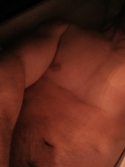 The image shows a close-up view of a person's bare torso, with warm lighting that casts a reddish hue over the skin. The person's left arm and shoulder are visible on the left side of the frame, and their chest is in the center. There is a dark background with some shadow play. The image does not contain any identifiable landmarks, people, or objects that would allow for a specific location within Lyuban, Russia, or any other city to be determined.