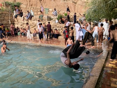 The image depicts an outdoor scene in Killi Malik Sher Mohammad, Pakistan, featuring a large gathering of approximately 30 to 40 males, predominantly boys and young men, around a rectangular, light blue-green water pool. The central action captures a boy mid-air, inverted, performing a backward flip or dive into the water, wearing a light-colored t-shirt and dark pants. Several other boys are partially submerged in the pool, with one near the left edge covering his mouth and nose. On the immediate tiled perimeter of the pool, multiple boys and young men stand, many with wet clothing, observing the activity. Beyond the pool's edge, ascending a terraced hillside, numerous individuals are seated on stone steps and ledges, forming an audience. Some clothing items are draped on the stone walls to dry. The background consists of rough-hewn stone retaining walls and steps leading up to an exposed, light brown earthen hillside. Sparse green vegetation is visible, including a prominent leafy bush on the right side of the frame. The scene is brightly lit by natural sunlight.