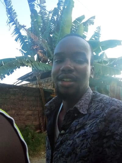 The scene is an outdoor daytime environment in Mtopanga ward, Kenya. A dark-skinned male, appearing to be an adult, occupies the foreground, facing the camera with an open-mouthed expression and a slight smile. He has short hair and a beard, wearing a dark, patterned button-up shirt open at the collar over a white undershirt. A partial view of another individual, exhibiting a white and neon-green striped garment on the shoulder, is visible on the far left. The background includes a weathered stone wall, dense banana trees with broad leaves, and a corrugated metal roof section. The sky is bright and clear above, suggesting natural daylight.