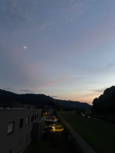 The image captures a twilight scene in Hausach, Germany, featuring a modern building in the foreground with a pathway and a grassy area to the right. The sky displays a gradient of blue and soft pink hues, with a sliver of the moon visible in the upper left quadrant. A dense forest line forms the backdrop, and a river runs parallel to the pathway. Distant structures, including what appears to be a tower on a hilltop, are subtly illuminated. The building in the foreground is multi-storied, with visible windows and architectural details. Streetlights are present along the pathway, casting a warm glow. The overall ambiance is serene and tranquil, indicative of the early evening hours.