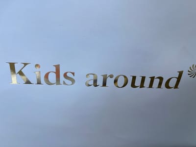 A metallic gold foil logo is emblazoned on a white surface. The text reads "Kids around". The font is elegant and serifed, with the letters slightly spaced apart. To the right of the text, a small, stylized pinwheel or sunburst graphic is visible, also in metallic gold. The background is a plain, light gray, suggesting a neutral backdrop or packaging. The lighting appears to be soft and even, casting minimal shadows, which could indicate an indoor setting or an overcast day. There are no people or other objects visible in the frame, focusing all attention on the text and graphic. The location context of Noventa di Piave, Italy, does not provide any direct visual cues within the image itself, as the scene is minimalist and lacks specific environmental details.