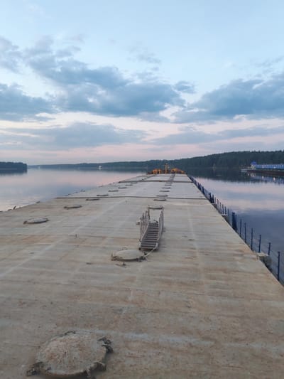 A low-angle, wide shot depicts a concrete pier extending into a body of water under a sky with blue and gray clouds.
The pier's surface is textured with circular hatches and a metal staircase. Along its right edge is a blue railing. At the pier's far end, several people wearing orange safety vests are visible.
Across the water, a shoreline is defined by dense trees. A building with blue roofing stands at the water's edge, its lights reflecting in the water. The body of water is calm with reflective qualities, mirroring the colors of the sky. The sky is a blend of light blue and gray, with hints of pink along the horizon. The location is in Ermolino, Russia.