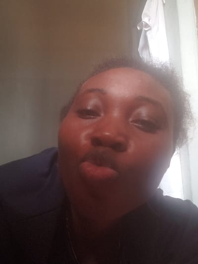 The scene is a close-up, front-facing self-portrait of a single individual in Agunmo, Nigeria. The subject has dark skin, dark, curly hair, and wears a dark garment with a visible thin, silver-colored necklace. Their eyes are partially closed, and lips are puckered. The background to the left is a plain, light brown wall. To the upper right, a white fabric item, possibly a shirt, hangs against a brighter, diffuse light source, consistent with a window. Lighting appears to originate from the right, creating a shadow on the subject's left side.
