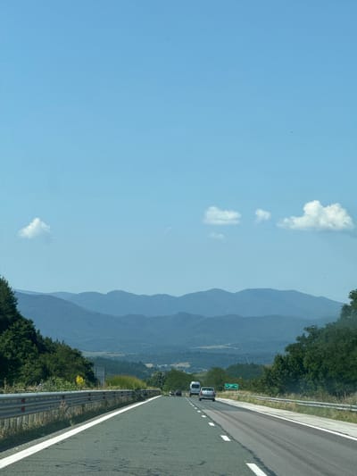 This image captures a view from a highway in Ravnishte, Bulgaria. The highway stretches forward, with white lines marking the lanes and a metal guardrail to the left. Green trees and vegetation line the side of the road, leading to rolling blue mountains in the distance under a clear blue sky with scattered white clouds. Several vehicles are visible on the highway, including a white van, a silver car, and other cars further ahead. A green road sign is also present. The overall scene depicts a serene journey through a scenic landscape.