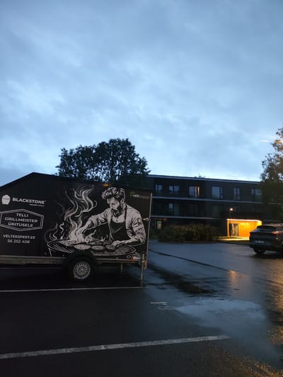 The image depicts a food trailer parked in a dimly lit outdoor area, likely a parking lot, in Tiskre, Estonia. The trailer is adorned with a large graphic featuring a man grilling burgers over flames, and text in Estonian that translates to "BLACKSTONE® CERAMIC GRILLS" and "TELLI GRILLMEISTER ÜRITUSELE" which means "ORDER GRILLMASTER FOR EVENT". Below this, "VELTEKSPERT.EE 56 252 408" is visible, indicating a website and phone number.

In the background, a modern, multi-story building with illuminated windows and an entrance glows with warm light. A dark-colored SUV is parked to the right of the building, its license plate reading "688 KVK". The ground is wet and reflective, suggesting recent rain. Trees and bushes are present, framing the scene. The sky is overcast and a pale blue, indicative of twilight or early morning. No people are directly visible, though the graphic implies human activity related to grilling. The overall atmosphere is somewhat moody due to the low light and wet conditions.