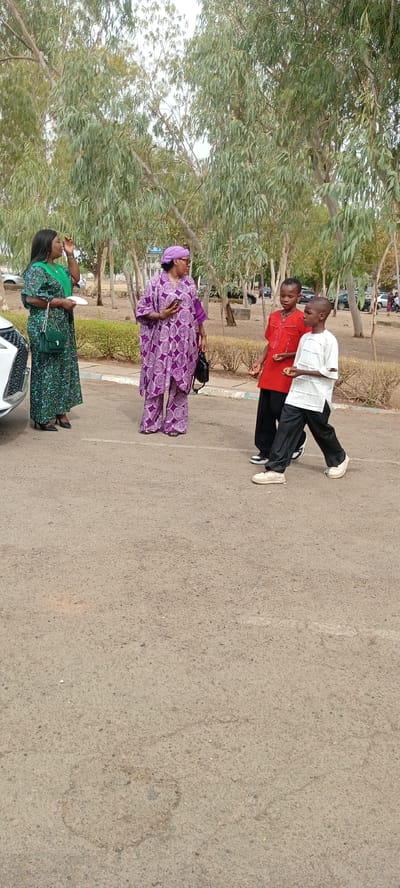 The scene is set outdoors on an asphalt surface, possibly a parking lot or road, in Goron Dutse, Nigeria. Three women and two boys are present. The woman on the left is wearing a long, green patterned dress and holding a white object. The woman in the center is wearing a purple patterned outfit with a matching head covering. She holds a phone in her right hand and a small black bag on her shoulder. The two boys are dressed in contrasting colors: one in a red shirt and black pants, and the other in a white shirt and black pants. The boy in the white shirt is walking toward the foreground. The trees with sparse leaves are visible in the background.