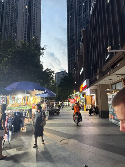 This media file captures a street scene in Haizhu District, China, likely during the late afternoon or early evening. The orientation of the image suggests a person is holding the camera and looking down at the scene. The setting is a bustling marketplace along a pedestrian walkway, with a large building on the left and shops lining the right side of the street.
Several people are visible, including a person on a scooter wearing an orange jacket, and shoppers browsing various stalls. One woman in a dark dress is prominently featured, looking towards the market. Another person, partially visible, is seen from behind, also near the market stalls. The stalls themselves are filled with clothing and other merchandise, displayed on racks and tables, with a large blue umbrella providing shade over some of the goods. Various motorcycles and scooters are parked along the walkway, indicating a common mode of transportation in the area.
The time of day appears to be transitioning from daylight to dusk, as indicated by the lighting and the sky, which shows a mix of clouds and a soft sky. The environment seems urban and active, with the architecture of the buildings suggesting a commercial or residential area. The presence of street vendors and a high volume of foot traffic points to a lively commercial district. There is some visible text on signs and banners, but it is mostly illegible in this resolution. The overall mood is one of everyday commerce and urban activity.