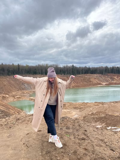 A woman stands at the edge of a steep, sandy embankment overlooking a body of water. She wears a beige trench coat, dark pants, white sneakers with red accents, and a purple beanie. Her arms are outstretched. The water is flanked by sandy cliffs, leading into a forest in the background. The sky is overcast with heavy clouds. The location is Ivlevo, Russia.