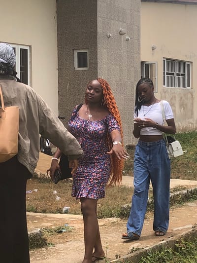 The image, taken in Farin Yashe, Nigeria, depicts three individuals outdoors. In the foreground, a person wearing a grey jacket over a black garment is partially visible, holding a mobile phone and a tan bag. Their hair is covered by a patterned headscarf. Behind them, a woman with long, orange curly hair is wearing a floral-patterned dress and is holding her left hand up, adorned with jewelry. Her face is turned downwards. Further back and to the left, another person in a white top and blue jeans is seen from the torso up. They have dark braided hair and a white purse slung over their shoulder. This person appears to be looking down. The scene is set in an outdoor environment with buildings and vegetation visible. The ground is a mix of dirt and some sparse grass, with small pieces of debris scattered around. The lighting suggests it is daytime, and the weather appears to be fair. There is no visible text in the image.
