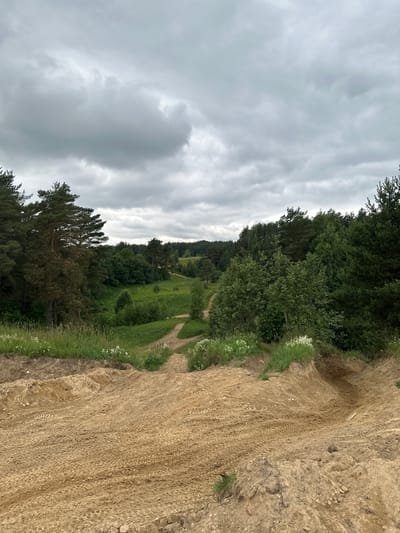 The image depicts a sandy track winding through a forested landscape under a cloudy sky. The foreground is dominated by the churned-up sand of the track, showing tire marks from vehicles. The track curves and descends into a wooded valley. The valley itself is filled with lush green grass and various trees, including pines and deciduous varieties. A winding path, likely an extension of the sandy track, is visible in the distance, leading further into the trees. The overall impression is of an off-road or motocross track located in a natural, somewhat remote environment, possibly within or near the city of Tokkari-Lend, Russia.