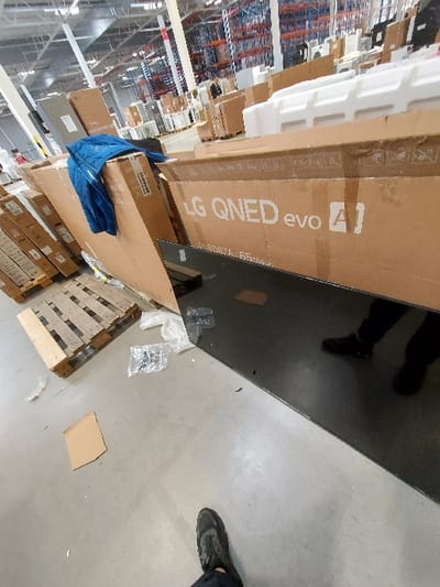 This image depicts an indoor warehouse or large retail space in Senec, Slovakia, characterized by a light grey concrete floor and high industrial ceilings with visible infrastructure. In the immediate foreground, a person's left foot, shod in a dark athletic shoe, is visible at the bottom-center. To the right, a large, reflective black surface, likely a television screen, lies horizontally, reflecting the lower legs and dark shoes of a person standing nearby. Adjacent to this surface on the floor are scattered pieces of clear plastic packaging and a small, rectangular piece of cardboard.

In the midground, a wooden pallet rests on the floor to the left. Numerous large cardboard boxes are present, some stacked. A prominent brown cardboard box in the center features the branding "LG QNED evo A]" and text indicating "ONED67A | 65 inches," identifying it as packaging for a 65-inch television. A blue puffer jacket is draped over a stack of cardboard boxes on the left. White foam packaging material is visible behind some of the boxes.

The background reveals extensive industrial shelving units stocked with additional boxed items, reinforcing the warehouse environment. A single individual is discernible standing in the distant background near these shelves. The overall scene suggests a process of unboxing or preparation of consumer electronics within a storage or distribution facility.