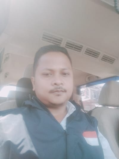 A man is captured in a selfie taken from inside a vehicle, likely a car, based on the visible interior elements like the ceiling vents, car seats, and window. The man, who appears to be of Southeast Asian descent, is wearing a dark blue jacket with a patch featuring the Indonesian flag (red and white horizontal stripes) on the left chest. He has short dark hair, a mustache, and is looking directly at the camera. The background outside the car window suggests an urban or semi-urban environment in Grong-Grong, Indonesia, with buildings and possibly some trees visible. The lighting is bright, indicating it's daytime. The image is slightly overexposed, particularly in the upper half.