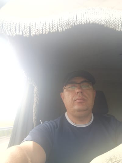 A selfie taken from the perspective of someone sitting inside a vehicle, likely a truck or van. The subject is a middle-aged man wearing a dark baseball cap and eyeglasses. He is looking directly at the camera with a neutral expression. The man is wearing a dark blue t-shirt with a lighter colored collar. The interior of the vehicle is visible, including a fringe-like decorative trim along the top of the windshield and a headrest of a seat behind him. Sunlight is streaming in from the left side of the frame, creating a bright glare and obscuring some details of the environment outside the vehicle. There is no information in the image to suggest the location in the city of Gusyne, Russia.