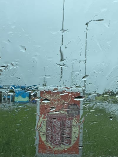 The image shows a view through a rain-streaked window of a Coca-Cola advertisement displayed outdoors. The advertisement is red and yellow, with what appears to be two soda bottles depicted on it. To the left of the advertisement, a blue and green structure is visible, possibly a building or a bus. In the background, the sky is overcast, and tall poles or masts are present, consistent with an outdoor advertising display or a sports field. The foreground is dominated by raindrops on the glass, distorting the view of the scene. The overall impression is of a gloomy, rainy day in Tiskre, Estonia, with the advertisement serving as the primary point of focus.
