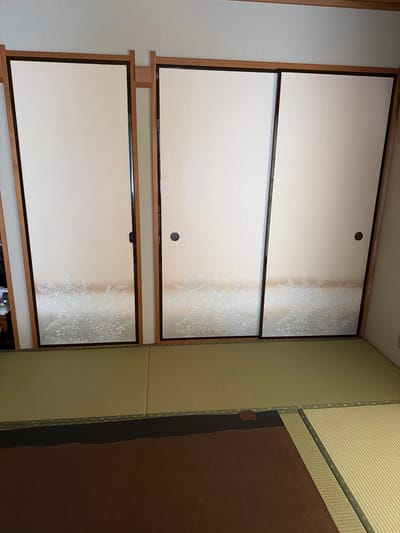 A room in Kawasaki, Japan, features three sliding paper doors (shoji) stacked vertically, framed in dark wood with light wood trim. The doors are mostly white with a patterned design along the right side. The room also contains tan tatami mats, and a brown section on the right.