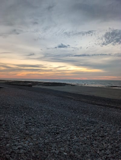 The image depicts a serene beach scene at dusk in Veules-les-Roses, France. The foreground is dominated by a vast expanse of dark, rounded pebbles. Beyond the pebble beach, a strip of sand meets the gentle waves of the English Channel. In the middle ground, the water is calm, with some rocky outcrops visible. Further out, the sea stretches towards the horizon, where it meets a sky filled with a tapestry of clouds. The sky displays a gradient of colors, transitioning from soft oranges and yellows near the horizon, indicative of the setting sun, to cooler blues and grays higher up. The clouds are textured and varied, with some appearing wispy and others more defined. Two small figures are visible in the distance on the wet sand near the water's edge, suggesting a moment of quiet contemplation or a leisurely stroll. The overall atmosphere is tranquil and contemplative, capturing the peaceful beauty of a coastal evening.
