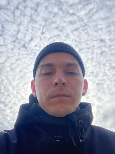 A low-angle, head-and-shoulders portrait of a male subject against a cloudy sky, reportedly taken in Nemchinovka, Russia. The individual, appearing to be an adult male, is centered in the frame, looking directly downwards towards the camera with a neutral expression. He is wearing a dark-colored knit beanie and a dark hooded jacket or sweatshirt. A white wireless earbud is visible in his right ear. His skin tone is fair, with slight redness observed on the nose and cheeks. The background consists entirely of a densely overcast sky featuring numerous small, scattered altocumulus clouds. No other environmental details are visible.