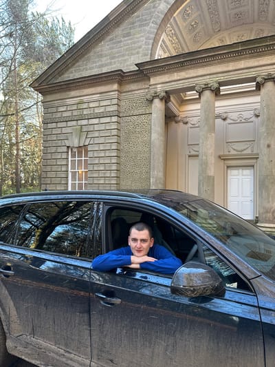A dark-colored SUV, visibly soiled with mud on its lower body panels, is positioned outdoors in front of a substantial stone building. A male individual is partially visible through the fully lowered driver's side window, leaning forward with his forearms resting on the window frame and hands interlocked. He has short dark hair, a light complexion, and is wearing a royal blue long-sleeved top. He maintains a direct gaze towards the camera with a neutral expression.
The background features a prominent classical-style stone building, identified as a section of Shugborough Hall, located near Stafford, United Kingdom. The building exhibits a triangular pediment atop an entablature, supported by at least two visible Corinthian columns forming a portico. Intricate friezes and ornate ceiling details are discernible within the portico. To the left of the columns, the building displays block-like ashlar stonework with a distinctive decorative chain-link pattern frieze and a textured stone panel. A rectangular window with a white sash frame is present on this section. A white double door is visible within the portico. Bare-branched trees are visible in the far left background under a bright sky.