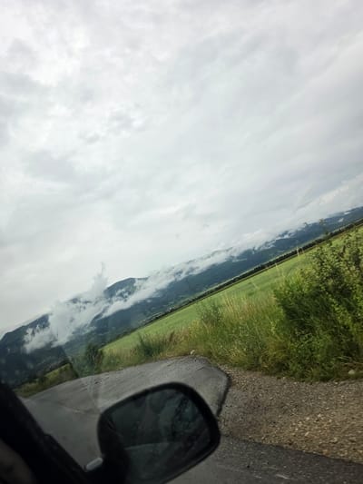 A tilted, eye-level, outdoor, daytime, full shot captures a view from a vehicle traveling along a paved road towards a lush green field and misty mountains. The road, wet and appearing to be asphalt with some gravel on the shoulder, curves slightly to the right. In the lower right corner, a black side-view mirror is visible, reflecting the overcast sky. Beyond the road, a vibrant green field stretches towards a range of dark green mountains partially obscured by wisps of white clouds. Tall, dry grass and green bushes line the edge of the field and road. The sky is filled with a dense layer of white and gray clouds, suggesting a potentially overcast or rainy day. The location is identified as being in Manolovo, Bulgaria.
