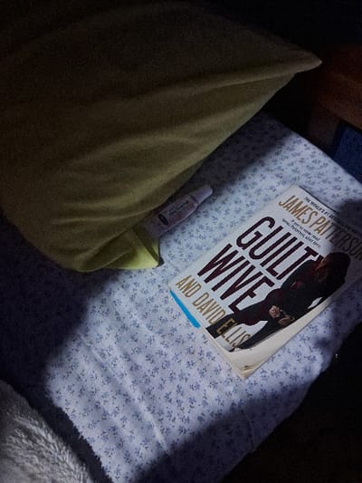 The image captures a dimly lit indoor scene, likely a bedside area, set in Muthambi ward, Kenya. A distinct rectangular beam of light illuminates parts of the bed, suggesting evening, night, or a specific lighting setup, creating strong contrasts between light and shadow.
The primary subject is a book titled "GUILT WIFE" (part of "Wife" is obscured, could be "Women"), co-authored by James Patterson and David Ellis. The book cover prominently displays "JAMES PATTERSON" with the descriptor "THE WORLD'S #1 BESTSELLING AUTHOR," and smaller text stating, "IF YOU'VE NEVER READ James Patterson, START HERE." The book is open and resting on a white sheet with a small, blue floral or patterned design.
Next to the book, tucked between it and a yellowish-green pillow, is a small, white plastic bottle or tube. Partially visible text on the bottle indicates "Body Butter" and "Rose & Lavender," along with a volume of "1 fl oz / 30 mL." The brand name is difficult to discern but might begin with "Good..." or "Gold...".
The pillow, made of a plain greenish-yellow fabric, occupies the left side of the frame. To the right, a fluffy, white blanket or throw is partially visible, adding texture to the scene. There is no direct activity depicted, but the arrangement of the book, pillow, and personal item suggests a quiet, domestic moment of rest, relaxation, or winding down, possibly before sleep or during a period of reading. The overall atmosphere is calm and peaceful.