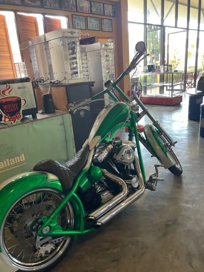 The image depicts the interior of a commercial establishment, likely in Thailand, given the partial visibility of "ridethailand.com" on a display surface. The primary subject is a custom chopper-style motorcycle, predominantly metallic green with white flame accents on the fuel tank and fenders, and numerous chrome components.

The motorcycle is parked on a light gray concrete floor, angled towards the right side of the frame, supported by its kickstand. Key features include extended chrome front forks, high-rise handlebars with a single circular mirror, a black quilted leather seat, a chrome "S&S Super" engine cover, and dual chrome exhaust pipes on the right side. Both front and rear wheels are spoked with disc brakes.

In the left background, behind the motorcycle's seat, a green display counter features a "WEST COAST Cafe" sign with a "Saeco" logo, alongside a black coffee grinder. Further left, several tall, light-colored display cases, some appearing to hold sunglasses, are visible. Above these, framed pictures are hung on an orange wall, with large wooden louvered shutters providing natural light from the far left.

The right side of the background is dominated by large, floor-to-ceiling glass windows and doors, offering a bright view of an outdoor area with greenery and a wooden bench. In front of these windows, a glass display table with a red base is present, along with a red portable fan on the floor. Two dark, barrel-shaped objects serve as potential tables or seating on the far right. A dark box branded "Jack Daniels Old No. 7" is positioned on a counter behind the motorcycle's handlebars. No individuals are present in the scene.