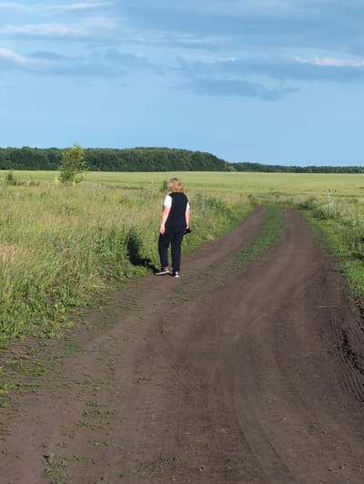 A woman wearing a black vest over a white t-shirt and black pants walks away from the camera on a dirt road. She has blonde hair. The dirt road is unpaved and appears to be a rural path, with tire tracks visible. To the left of the road is a grassy field with tall green and yellow vegetation and some small bushes. In the background, there is a dark green line of trees forming a treeline, and beyond that, a bright blue sky with scattered white clouds. The location is described as a field in the city of Вышки, Russia, likely a rural or natural area on the outskirts.