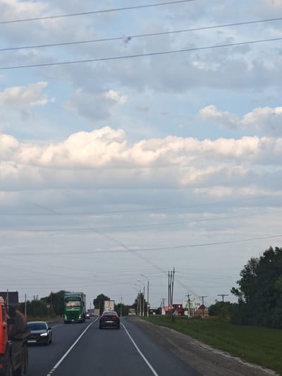 This image depicts a highway scene in the vicinity of Kizhevatovo, Russia. The sky is filled with scattered clouds, with power lines crisscrossing the upper portion of the frame. The road itself is asphalt, with white lane markings.
In the foreground, a large orange truck is partially visible on the left side, suggesting it is also traveling on the highway or parked nearby. A black Opel Astra is in the left lane, with the license plate visible. In the right lane, a black Audi is traveling ahead.
Further up the road, a bright green semi-truck, likely a Volvo, is in the left lane. Several other vehicles, including a white truck and what appears to be a small bus or van, are visible in the distance, indicating traffic flow.
The right side of the highway features a grassy shoulder, a dense line of trees, and some buildings in the distance, some of which appear to be houses or small commercial structures. Several utility poles and signs are also present along the roadside. One sign appears to indicate "5 KM," possibly a distance marker. The overall impression is a typical rural or suburban highway environment.