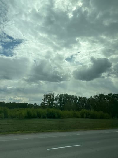 A landscape scene showing a road running alongside a green field with a line of trees separating them. The sky is filled with clouds, some darker than others, with a hint of blue sky peeking through. The image is likely taken during the day. There are no people visible in the scene. The road is gray and has a white line marking the edge. The scene appears to be in a rural or semi-rural setting. While the prompt requests that this be determined to be in Pokrovka, Russia, there is not sufficient information here to determine a location from the image.