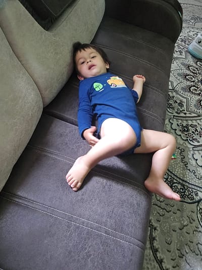 A young child, likely a toddler, lies on their back on a dark gray sofa. The child is wearing a blue long-sleeved bodysuit with a green leaf design on the left chest and a cartoonish yellow vehicle on the right. The child's legs are bent, and their feet are visible. The sofa has white stitching detailing its seams. In the background, a patterned rug with intricate designs in light gray and beige is visible on the floor. The lighting suggests it is daytime, possibly indoors. The child appears relaxed, with their eyes open and mouth slightly ajar, looking upwards. The scene is set in a domestic environment, possibly a living room, in Yakkabog, Uzbekistan. No visible text is present in the image.