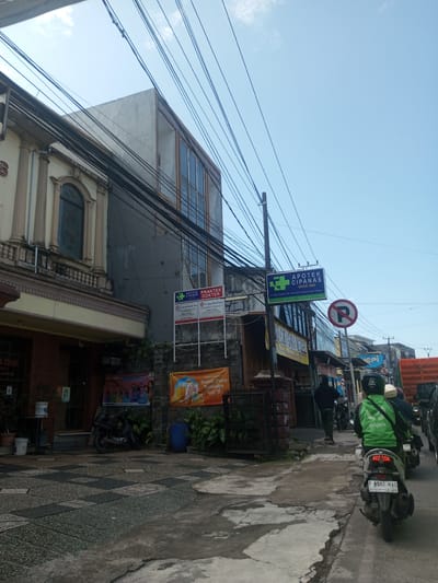 The image shows a street scene in Cipanas, Indonesia, with buildings on the left side of the frame. The architectural styles of the buildings are diverse, including a European-style building with ornate detailing. Next to it is a modern building with a grey exterior. There are signs visible on the buildings, including one for Apotek Cipanas, a pharmacy established in 1982. There is also a sign indicating a medical practice with the name "Dr. Sagung Hindi Procto." On the street, there is a 'no parking' sign and several parked motorcycles. In the foreground, a Gojek motorcycle is stopped in traffic, and a person wearing a Gojek jacket is sitting on it. Other vehicles and pedestrians are also present. The street surface is cracked and uneven. Overhead, there are numerous power lines. The sky is light blue with some clouds. The location is identified by the Apotek Cipanas sign with the text "Jl. Raya Cipanas No. 144, Kec. Cipanas, Kab. Cianjur."