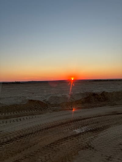 This is a vertical, wide-angle photograph of a desert landscape at sunset. The sun, a bright orange orb, is low on the horizon, casting a warm glow across the scene. Its rays create a lens flare effect, emanating outwards and illuminating the dusty ground. The sky transitions from a pale blue on the left to a gradient of orange and yellow as it meets the horizon.

The foreground is dominated by a rough, uneven surface of dry earth and sand, marked with deep tire tracks, suggesting recent vehicular activity. This textured terrain occupies the right half of the image. In the distance, a faint, dark line of vegetation or structures can be seen along the horizon, almost blending into the sunset's haze.

The overall mood is serene and slightly desolate, characteristic of a desert at dusk. The image captures the natural beauty of the transition from day to night in an arid environment. No people, objects, or text are explicitly visible. The time of day is clearly sunset, indicated by the sun's position and the sky's colors. The location cues suggest a remote, undeveloped area, possibly a construction site or a natural desert terrain.