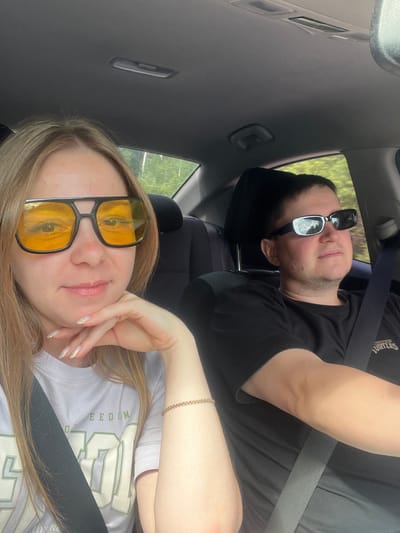 Two people are inside a car, with the camera positioned to capture a selfie-style shot. The woman in the foreground is wearing large, yellow-tinted sunglasses and a white t-shirt with green lettering. Her left hand is resting on her chin, and she has long blonde hair. The man is in the background, wearing dark sunglasses and a black t-shirt. He is looking slightly to the right. Both individuals appear to be wearing seatbelts. The interior of the car is visible, including the ceiling and part of the dashboard. Outside the car, green trees and foliage can be seen through the windows, suggesting a rural or suburban setting. The image was taken during the daytime.
