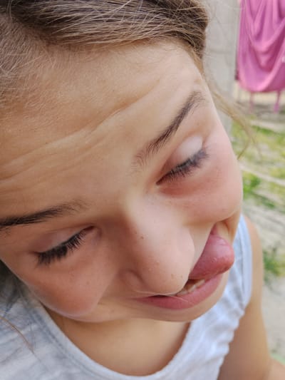 The media file is a close-up, upward-angled shot of a young person's face. The subject, likely a child, is sticking their tongue out and making a playful, silly expression. Their eyes are closed, and their eyebrows are furrowed, adding to the comical look. Freckles are visible on the bridge of their nose and cheeks. The person is wearing a white tank top with a textured neckline.
The background is blurred but shows hints of an outdoor setting with green foliage and what appears to be a pink draped fabric hanging in the distance. The lighting suggests it is daytime, possibly a bright day given the exposure. The scene is candid and captures a moment of lightheartedness. No text is visible in the image. The location context provided is Vulcănești, Moldova.